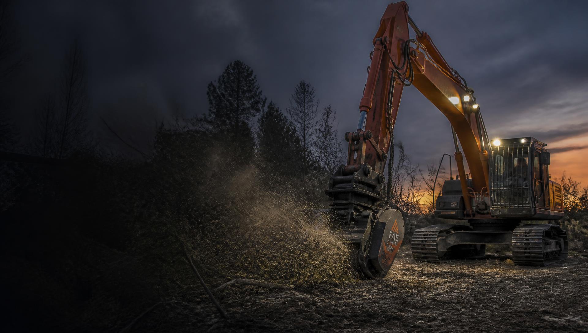 Excavator with a forestry mulching head clearing brush at dusk